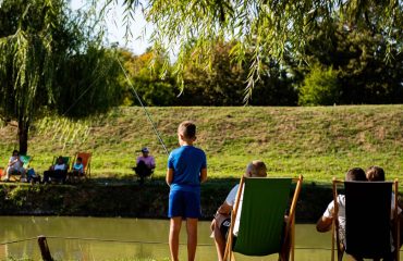 Young boy fishing by a pond on a sunny day, surrounded by trees and relaxed people.